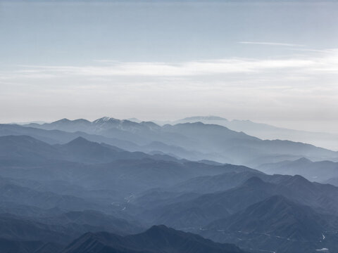 Aerial view of layered blue-gray mountain ridges fading into haze, distant peaks dusted with snow under a soft sky near Santa Clarita, California