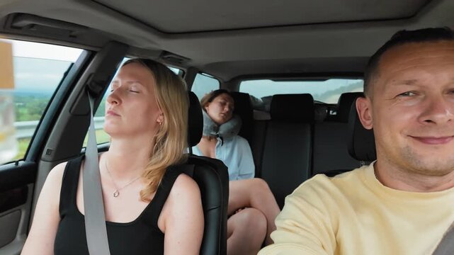 Positive man driving a modern car on a highway during a long road trip while his wife and teenage daughter are sleeping in the passenger seats, illustrating a family travel concept