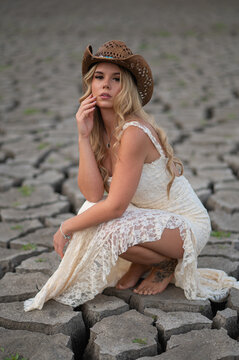 Blonde woman in cowboy hat crouching on dry cracked ground