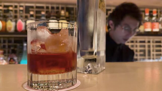 Close up of cinematic negroni cocktail with ice on bar counter with bartender working in background.