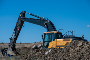 A heavy yellow and black crawler excavator works behind a large mound of dug earth with its boom arm curved against a solid blue sky. © Andrey