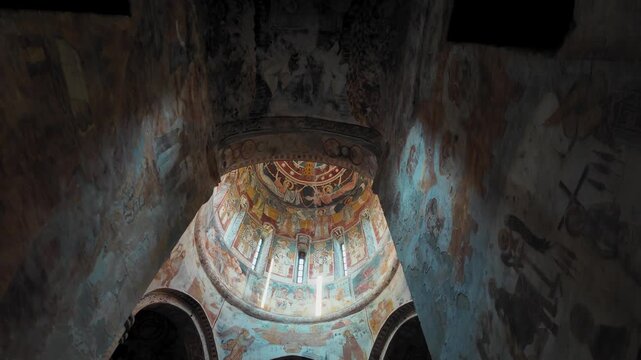 camera slowly moves forward and tilts upward inside old Orthodox church of Nikortsminda in Georgia, revealing majestic dome with medieval frescoes and sunrays breaking through narrow windows of drum