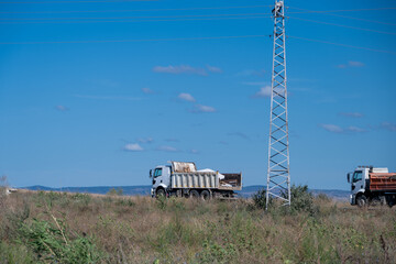 Heavy-duty dump trucks drive across an open rural worksite near a tall steel transmission tower under a vivid blue sky. © Andrey