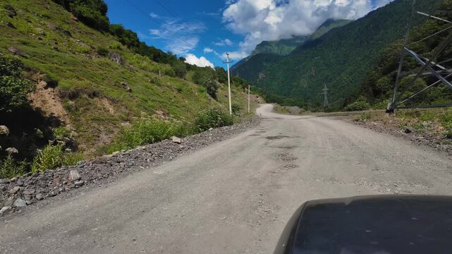 Pov footage from a vehicle traveling on a rough, unpaved country road through a scenic mountain valley on a sunny day with blue sky and clouds, showcasing a beautiful summer landscape