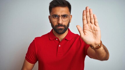 Fototapeta premium Young bearded man in red polo shirt makes stop gesture with open palm. Serious male looking at camera, raising hand to signal refusal, warning prohibition. Caucasian adult shows rejection,