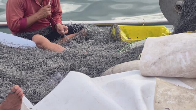 Central Vietnamese fisherman sitting on a wooden boat mending and untangling a large fishing net at a coastal port