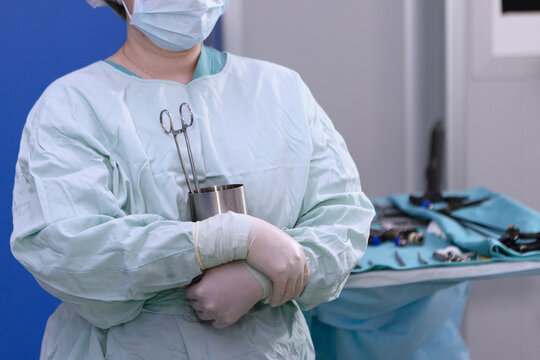 Nurse in green lab coat stands with her arms crossed over her chest. Blurred background of an operating room with instruments. She holds stainless steel cup with surgical instruments in her hands