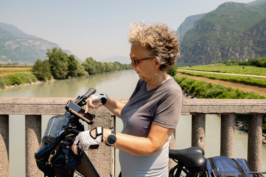 Senior woman checking phone by Adige River on bike trip