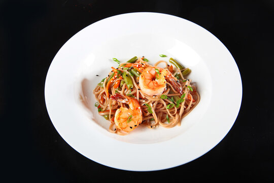 Asian noodles with shrimp and vegetables in white plate. Asian stir fried noodles with shrimp, green beans, carrots, sesame seeds and green onions in white bowl. Dark background