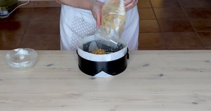 Crushed Biscuits with Melted Butter Placed into a Cheesecake Mold. Close-up of female hands transferring crushed biscuits mixed with melted butter into a metal cake mold lined with parchment paper.
