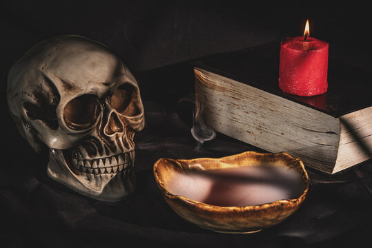 Human skull with burning candle, ancient book and mysterious red potion in smoking bowl