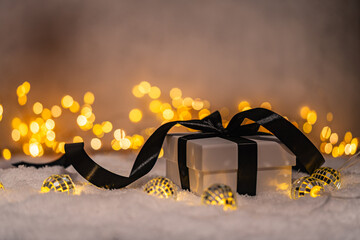 Naklejka premium A Christmas background featuring a white gift box tied with a black satin bow surrounded by black baubles, a teal star, and frosted pine on snow, lit by warm twinkling lights with copy space.