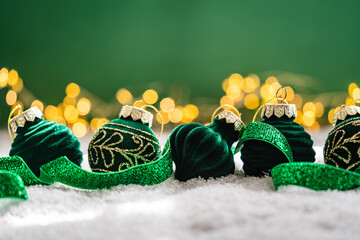 Naklejka premium A Christmas background featuring plush emerald velvet baubles with golden filigree detail and a glittering green ribbon on snow, set against deep green with twinkling lights and copy space.
