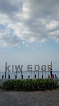 Large metal KIW EDGE sign standing on a waterfront promenade under a cloudy sky