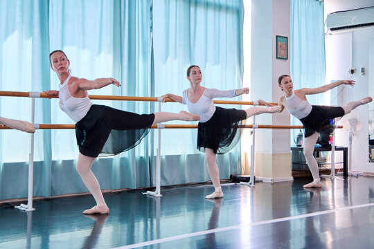 Adult female ballet dancers practice barre exercises in bright studio light
