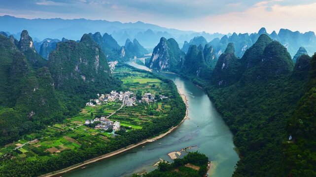 High angle view of the Li River winding past green karst mountain peaks, small village, and farmland under bright sunlight in Guilin, China.