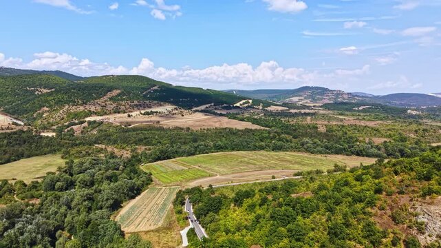 A wide view shows fields and hills in a valley. Green trees border the fields, and distant mountains rise under a blue sky with clouds. It is a clear day.