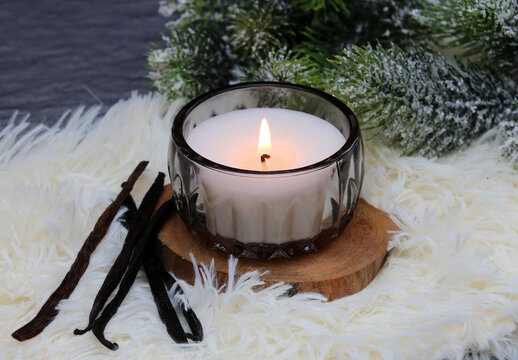 Cozy winter decoration featuring a burning white scented candle in a glass jar, vanilla beans, and snow-covered fir branches.