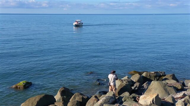 Small motorboat cruising offshore, wide view from a rocky coast with a person near the water, showing coastal transport, marine recreation, tourism activity and local boating along the Black Sea