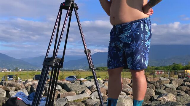 Man adjusting camera low angle on tripod outdoors near the coast, showing professional filming setup for media production, travel content, documentary work and commercial photography operations