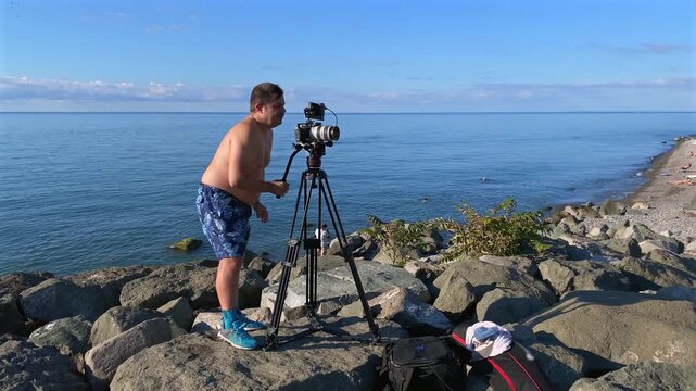 Producer filming coastal scene wide shot with professional camera on tripod, showing location production workflow for documentary, travel media, commercial content, field directing and outdoor shoot