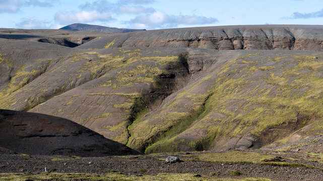 Barren volcanic slopes streaked with moss and eroded ravines create a dramatic terrain in the Icelandic highlands near Kerlingarfjoll under a partly cloudy sky.