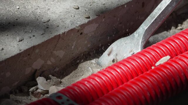 Pneumatic chisel breaking concrete beside vibrant red cable conduits at active work site. Close-up tracking shot.