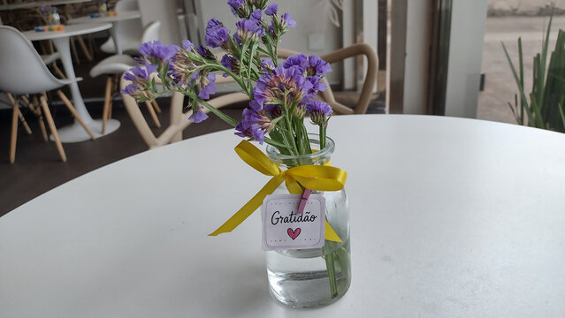 Small glass jar filled with water holding delicate purple statice flowers decorated with a bright yellow satin bow on a bright white cafe table.