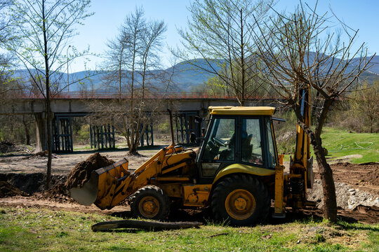A heavy yellow backhoe loader machine is actively operating on a grassy riverbank moving thick dark soil for a rural landscaping project with an old metal bridge standing in the background.