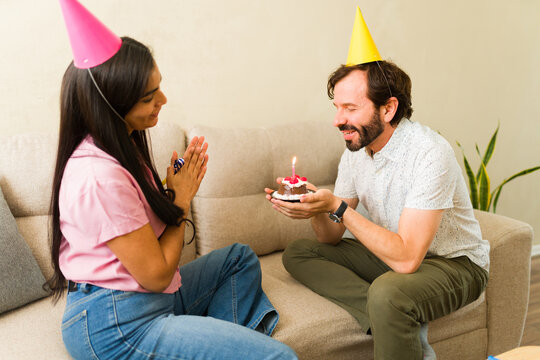 Couple celebrating birthday at home, man presenting small cake with candle to happy woman on sofa