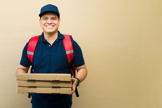 Latin delivery person holding a stack of three pizza boxes, wearing a uniform, cap, and red thermal backpack