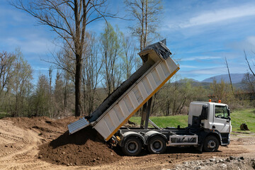 A heavy commercial dump truck lifting its large cargo bed to unload a massive quantity of dark fresh soil onto an outdoor development site surrounded by bare trees during a bright spring morning. © Christos