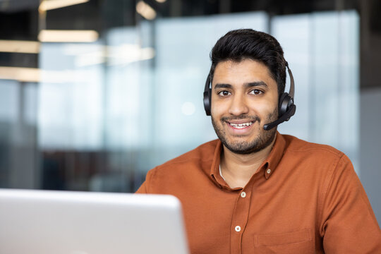 Smiling young man wearing headset sits at laptop in bright modern office, suggesting remote customer support, online assistance, professional communication, and friendly service in corporate workspace