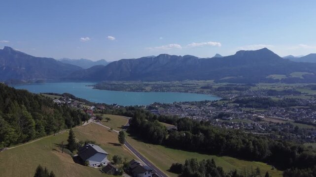Panoramic aerial view over alpine hillsides showing a winding road, rural houses, and the vibrant turquoise Lake Mondsee bordered by majestic mountain peaks under a clear sky.