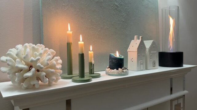 Horizontal 4K video showing a static low angle medium shot of a white wooden fireplace mantel decor. Features burning taper candles, white coral, ceramic houses, and a bioethanol fireplace.