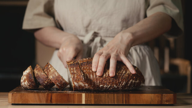 Close-up of baker in rustic linen apron slicing freshly baked artisan sourdough bread with knife on wooden cutting board. Homemade crusty loaf for breakfast, bakery promos, and healthy food concepts.