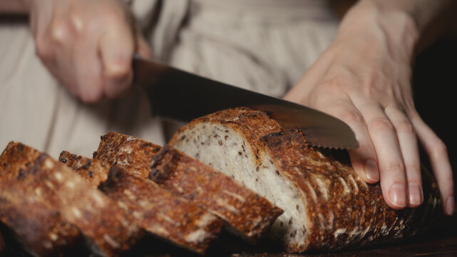 Close-up of baker in rustic linen apron slicing freshly baked artisan sourdough bread with knife on wooden cutting board. Homemade crusty loaf for breakfast, bakery promos, and healthy food concepts.
