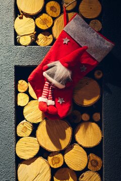 A vertical photograph of a red plush gnome Christmas stocking hanging on a recessed decorative wall filled with stacked wood logs