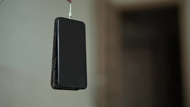 Broken black smartphone with detached screen hanging from cable, slowly spinning. Concept of internal parts, repair, e-waste, technology breakdown, planned obsolescence. Blurry background, nobody.
