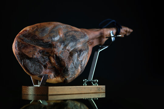 Side view of cured ham leg resting on wooden board. Hamon on professional jamon stand on black background. Dried ham on wooden stand with black cord on dark background