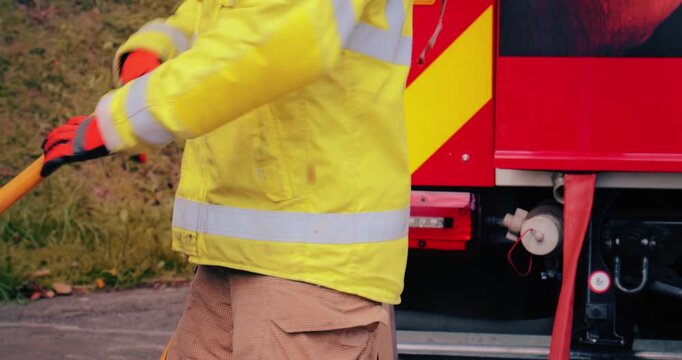 Firefighter team - fireman equipped with emergency services response firefighting crew uniform and gear reeling in fire hose. Department crew from fire station in scene background.