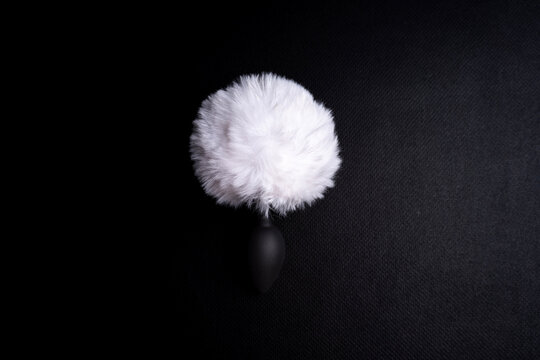 Black metal intimate plug with white fluffy faux fur bunny tail attached placed vertically on dark textured background.