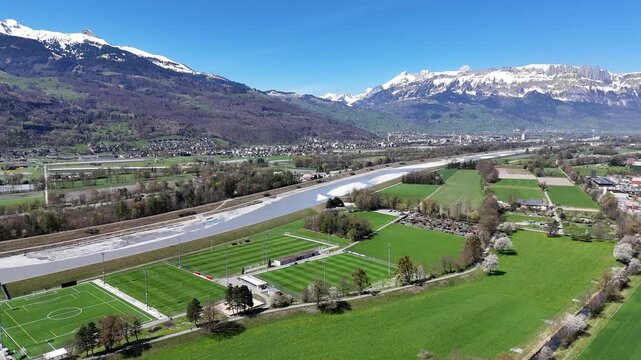rhine river valley aerial drone footage snow capped alps mountain backdrop wide gravel riverbed sports complex football pitches running track green farmland liechtenstein switzerland border landscape