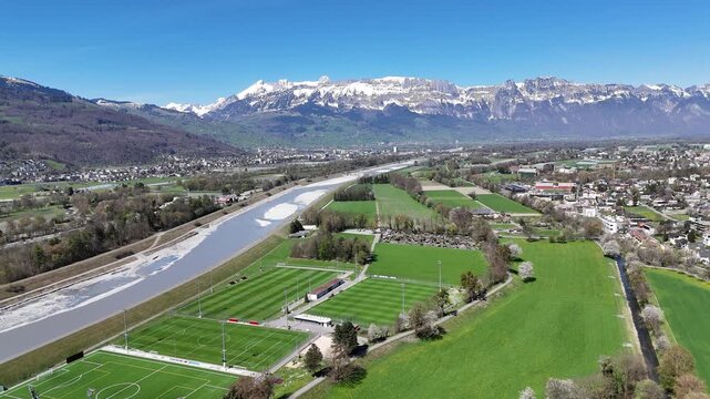 alps snow capped peaks towering rhine valley aerial view sports fields football pitches river gravel bars green meadows farmland alpine village rooftops liechtenstein switzerland border scenery