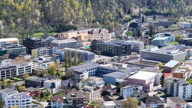 st florin cathedral spire vaduz town center aerial view government house town hall complex modern office buildings forested hillside rock face backdrop civic district liechtenstein drone footage