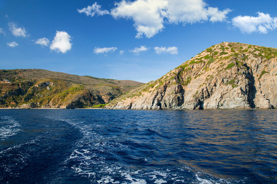 View of a Mediterranean island coastline with green hills and slopes from the sea under a blue sky with clouds.