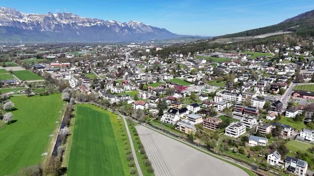 farmland fields alpine village rooftops snowcapped mountain range backdrop tree lined road spring blossom liechtenstein switzerland border drone footage aerial view establishing shot rural landscape