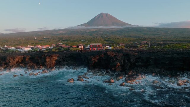Aerial view of a coastal village on Pico Island, Azores, featuring traditional houses on rugged volcanic cliffs, with the iconic Mount Pico volcano and a visible moon in the clear sky.