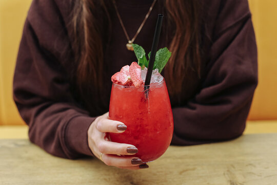 Woman holding red cocktail with ice background. Colorful chilled drink in rounded glass garnished with mint and fruit pieces. Refreshment alcohol beverage nightlife leisure party and social drinking.