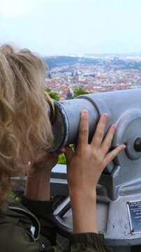 Blonde woman looks through coin operated telescope at city skyline. She steadies gray viewing scope with both hands while scanning distant rooftops from overlook, standing beside railing outdoors.